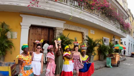 Un-Grupo-De-Turistas-Y-Varias-Palenqueras-Con-Vestidos-Coloridos-Balancean-Fruteros-Sobre-Sus-Cabezas-En-Una-Calle-Del-Casco-Antiguo-De-Cartagena-De-Indias,-Colombia.