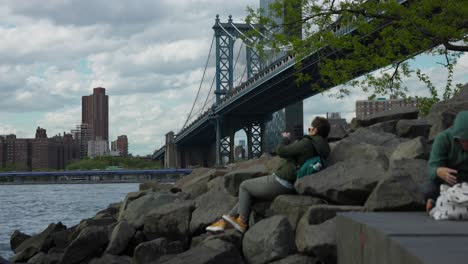 Tourist-Seen-Laying-On-Rocks-Beside-East-River-Taking-Photos-Of-Manhattan-Bridge-With-NYC-Skyline