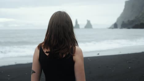 Young-beautiful-woman-in-black-dress-standing-on-black-sand-beach-Iceland,-looking-at-dramatic-waves-seascape,-slow-motion