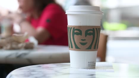 Auf-Einem-Tisch-Im-Amerikanischen-Multinationalen-Kaffeemarkengeschäft-Ist-Ein-Starbucks-Kaffee-Pappbecher-Zu-Sehen,-Während-Im-Hintergrund-Die-Kunden-Unscharf-Zu-Sehen-Sind