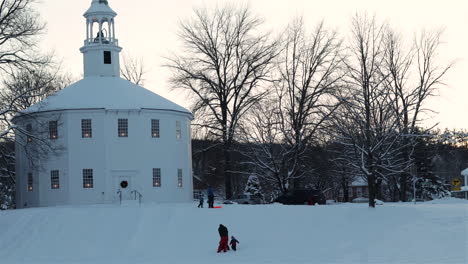 Time-lapse-of-children-and-adults-sledding-in-the-glow-of-evening-sunset-in-winter-next-to-The-Old-Round-Church-in-Richmond,-Vermont