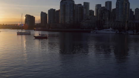 A-DOLLY-SHOT-Moving-Along-the-Rocks-to-Reveal-Vancouver's-City-Skyline-Illuminated-by-the-Evening-Light,-Boats-Float-in-the-Distance