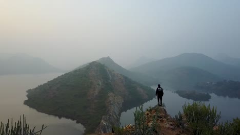 Hombre-Parado-En-El-Borde-De-Un-Acantilado-Rocoso-Y-Admirando-El-Paisaje-Brumoso-Del-Lago-Badi-En-Udaipur,-Rajasthan,-India
