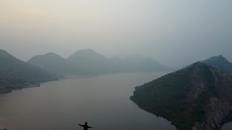 Impresionante-Paisaje-De-Las-Montañas-Costeras-Y-Aguas-Tranquilas-En-El-Lago-Badi-Con-Un-Hombre-Parado-En-El-Acantilado-En-Udaipur,-Rajasthan,-India