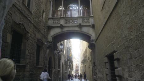 Pont-Del-Bisbe,-Barcelona,-Con-Pareja-Y-Turistas-Caminando-Por-Una-Calle-Estrecha-En-El-Barrio-Gótico-De-Barri