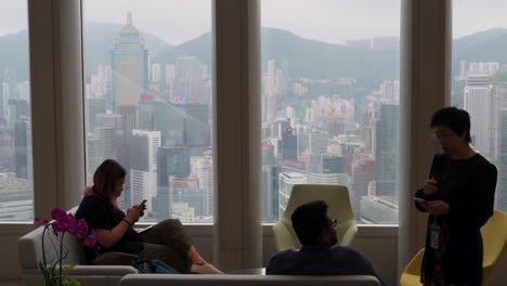 Menschen-Sitzen-Auf-Modernen-Möbeln-In-Einem-Hochhaus,-Während-Im-Hintergrund-Ein-Blick-Auf-Die-Skyline-Von-Hongkong-Zu-Sehen-Ist