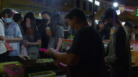 Chefs-Prepare-Food,-People-Walking-Past-Buying-Food,-Local-Night-Market,-Kuala-Lumpur,-Malaysia