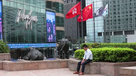 A-man-rests-outside-the-Exchange-Square,-the-building-housing-the-stock-market,-as-the-People's-Republic-of-China-and-Hong-Kong-SAR-flags-wave-in-the-wind-in-the-background