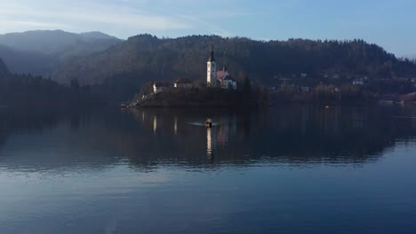 Aerial-Approaching-Church-Of-The-Mother-On-Bled-Island-On-Lake-Bled-Flying-Over-Passing-Boat
