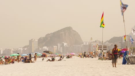 Statische-Weitwinkelaufnahme-Vieler-Menschen,-Die-Sich-An-Einem-Nebligen-Und-Windigen-Tag-Am-Sandstrand-Von-Rio-De-Janeiro-Entspannen