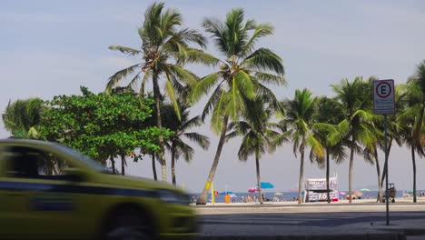 Wide-shot-showing-walking-people-along-road-in-front-of-Palm-trees-and-beach-In-Rio-de-Janeiro---Traffic-in-foreground