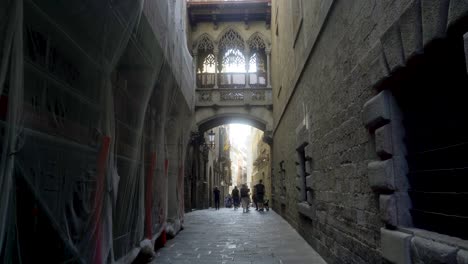 Pont-Del-Bisbe,-Barcelona,-Mit-Paar-Und-Touristen,-Die-Durch-Die-Enge-Straße-Im-Gotischen-Viertel-Barri-Gehen