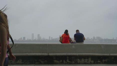 Couple-Sitting-On-The-Embankment-And-Watching-The-Back-Bay-And-Cityscape-During-The-Coronavirus-Pandemic-In-Mumbai,-India