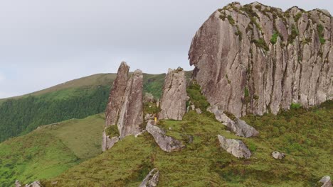 Orbit-shot-of-big-rock-formation-man-standing-in-yellow-jacket-at-Flores-island-Azores,-aerial