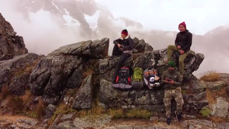 Dronie-Shot-Of-Three-Hikers-Resting-On-Hill,-Snowy-Mountains-In-Background,-Peru