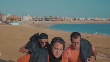 Ein-Hijabi-Athlet-Mit-Ein-Paar-Männlichen-Athleten,-Die-Mit-Hölzern-Und-Sandsäcken-Am-Strand-Entlangrennen-–-Zeitlupe