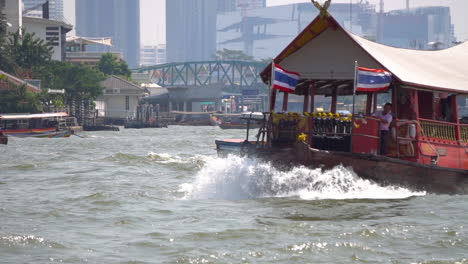 Vista-En-Cámara-Lenta-De-Un-Barco-Con-Banderas-Tailandesas-Navegando-Por-El-Río-Chao-Phraya-Con-El-Paseo-Marítimo-De-Bangkok-Al-Fondo