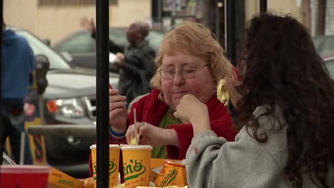 Zwei-Erwachsene-Frauen-Essen-Draußen-Fast-Food-Und-Sitzen-Am-Tisch,-Im-Hintergrund-Vorbeiziehende-Menschen-Und-Verkehr