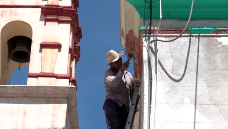 Man-with-hat-scraping-paint-off-the-wall-of-a-church-using-tall-ladder,-Long-shot