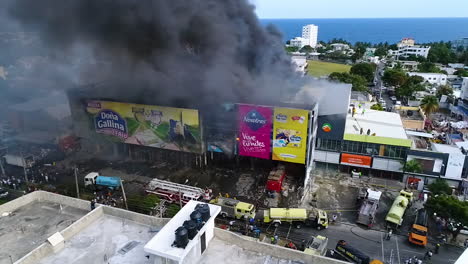 Aerial,-static,-drone-shot-of-firefighter-trucks,-in-front-of-a-warehouse,-on-fire,-dark-smoke-rising,-in-Santo-Domingo-city,-Dominican-Republic