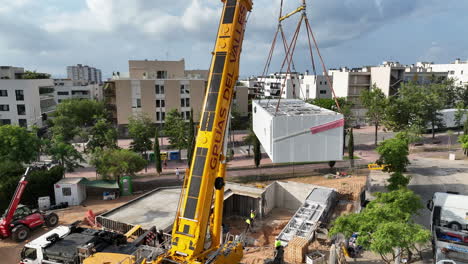 Aerial-view-circling-crane-lifting-smart-modular-housing-project-onto-construction-site-foundation