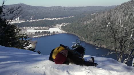 Caucasian-female-hiker-resting-in-the-snow-on-top-of-a-mountain-with-view-on-Longemer-lake,-Vosges,-France