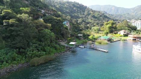 Drone-view-in-Guatemala-flying-over-a-blue-lake-next-to-a-green-mountain-with-a-wooden-walkway-and-a-hotel-on-a-hill-on-a-sunny-day-in-Atitlan