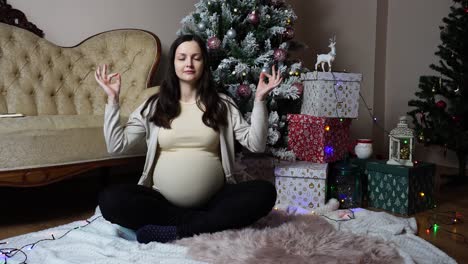 Pregnant-female-with-round-belly-sit-in-yoga-meditation-pose-near-Christmas-tree