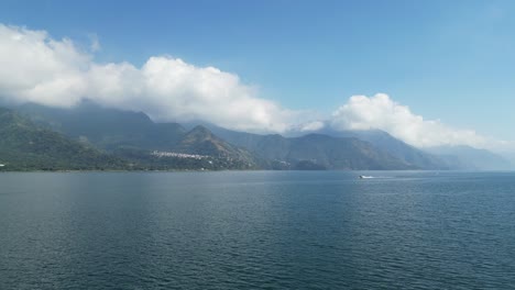 Drone-view-in-Guatemala-flying-over-a-blue-lake-surrounded-by-green-mountains-approaching-a-moving-boat-on-a-sunny-day-in-Atitlan