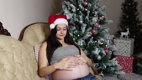 Pregnant-women-with-round-naked-belly-put-Santa-hat-on-head-near-Christmas-tree