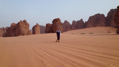 Hombre-Caminando-En-El-Desierto-Del-Sahara-Con-Una-Gran-Formación-De-Arenisca-En-El-Fondo-En-El-Parque-Nacional-Tassili-N&#39;ajjer,-Argelia