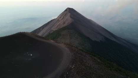 Vista-De-Drones-En-Guatemala-Volando-Sobre-El-Cráter-De-Un-Volcán-Cubierto-De-Cenizas-Al-Amanecer-Rodeado-De-Montañas-Verdes-Y-Nubes-Sobre-Ciudades