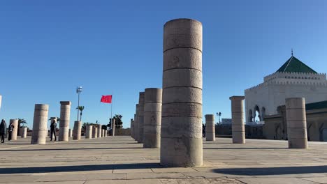 Antiguas-Columnas-De-Arenisca-Y-El-Mausoleo-De-Mohamed-V-Con-Bandera-Marroquí-Ondeando-En-El-Fondo