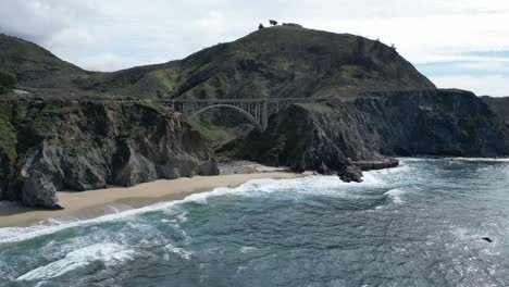 Drone-flies-over-the-water-showing-a-beautiful-beach-beneath-one-of-the-iconic-bridges-along-highway-101-near-Big-Sur,-California