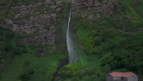 Rückwärtsfliegen-Am-Wasserfall-Mit-Lokalem-Haus-In-Flores,-Azoren---Drohnenaufnahme