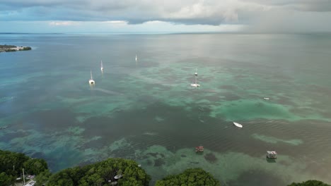 Drone-view-in-Belize-flying-over-caribbean-dark-and-light-blue-sea-with-boats,-a-white-sand-caye-covered-with-palm-trees-on-a-cloudy-day