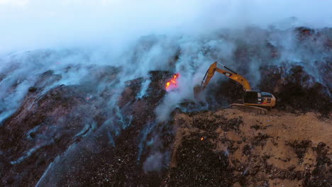 Aerial-view-of-the-firemen-using-a-Digger-to-extinguish-a-wildfire,-blue-smoke-rising-above-damaged-nature,-cloudy-evening,-in-Dominican-Republic---Dolly,-drone-shot