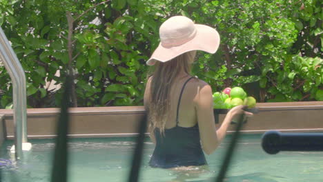 A-woman-in-a-black-bathing-suit-and-a-straw-hat-carrying-a-basket-of-fruit-over-her-private-swimming-pool