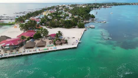 Drohnenansicht-In-Belize,-Die-An-Einem-Bewölkten-Tag-über-Das-Grüne-Und-Blaue-Meer-Der-Karibik,-Einen-Weißen-Sandcaye-Mit-Palmen-Und-Restaurants-Fliegt,-Während-Ein-Boot-Einen-Kanal-überquert
