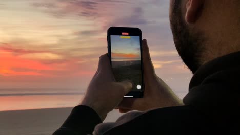 Junger-Männlicher-Tourist-Dreht-Video-Vom-Sonnenuntergang-Am-Strand-Auf-Modernem-Smartphone