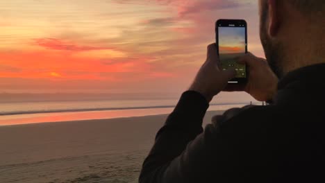 Young-man-male-tourist-takes-pictures,-shoots-video-of-sunset-on-beach-on-modern-smartphone