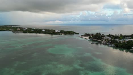 Drone-view-in-Belize-flying-over-caribbean-dark-and-light-blue-sea,-a-white-sand-caye-covered-with-palm-trees-and-restaurants-on-a-cloudy-day