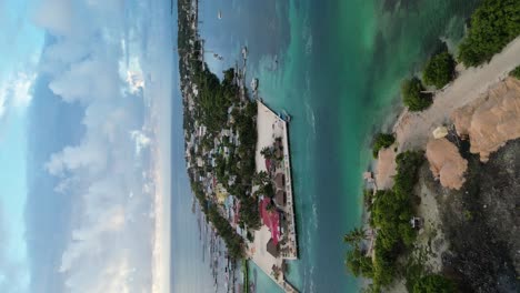 Drone-portrait-view-in-Belize-flying-over-caribbean-dark-and-light-blue-sea,-a-white-sand-caye-covered-with-palm-trees-and-restaurants-on-a-cloudy-day