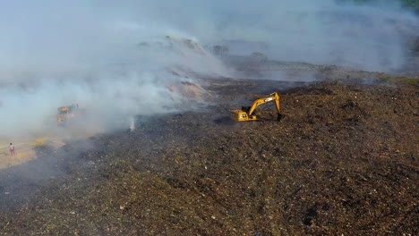 Aerial,-tracking,-drone-shot-of-the-fire-department-using-a-Excavator-to-extinguish-a-wildfire,-smoke-rising-in-destroyed-nature,-sunny-day,-in-Dominican-Republic
