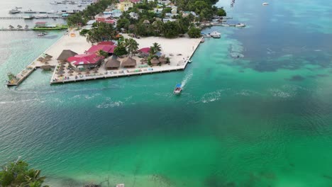 Drone-view-in-Belize-flying-over-caribbean-green-and-blue-sea,-a-white-sand-caye-with-palm-trees-and-restaurants-with-a-boat-crossing-a-canal-on-a-cloudy-day