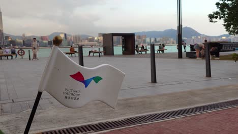 Plastic-Victoria-Harbour-Flag-With-Wan-Chai-Temporary-Promenade-In-Background-And-People-Walking-Past-In-Hong-Kong