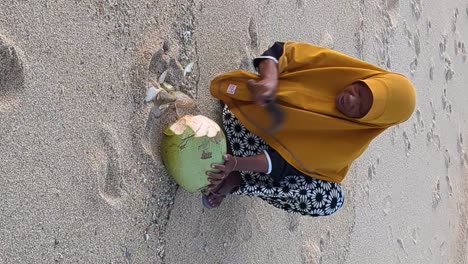 Local-lombok-woman-opening-a-coconut-for-a-tourists-on-the-beach,-vertical