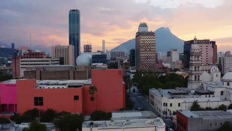 Ascending-drone-reveals-downtown-Monterrey,-Mexico-and-mountains-in-the-background-with-stunning-purple-orange-sunset
