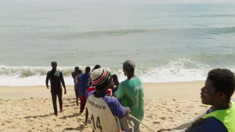 Los-Hombres-Levantan-La-Red-De-Pesca-Con-Una-Cuerda-Hasta-La-Playa-De-Arena-En-Ghana,-Una-Mujer-Pasa