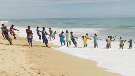Young-men-pull-fishing-net-from-ocean-to-sand-beach-in-Ghana,-wide